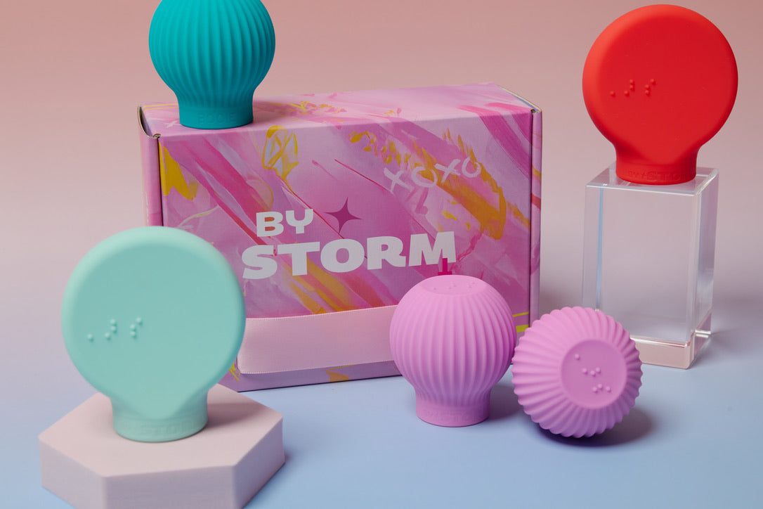 Four adaptive beauty grips in aqua, teal, red, and purple displayed around a pink and yellow ByStorm box with the logo visible. The products include flat circular grips and ribbed spherical grips, arranged on pastel surfaces with a pink to blue gradient background