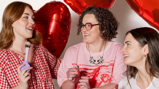 Three femme-presenting people smile and laugh together in a studio setting, surrounded by large red love heart balloons. 