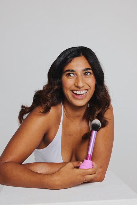 A woman sitting at a table, smiling, holding a makeup brush fitted with a pink ByStorm grip