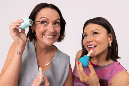 Two women smile and apply makeup using ByStorms Celeste Barber & Storm’s Accessible Beauty Tools That Are Changing the Game, celebrating inclusive beauty with innovative, adaptive designs against a plain light background.
