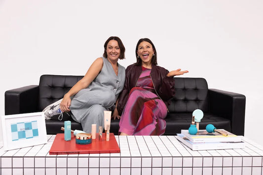 Two women smile on a black couch before a table with ByStorm beauty tools—backed by Celeste Barber—and books, highlighting the vibrant energy of women-led startups and their bold approach to accessible beauty.