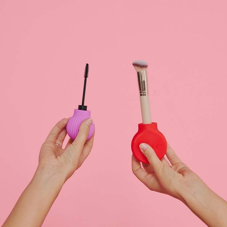 Two hands hold ByStorm’s Better Together Pack adaptive beauty tools against a pink backdrop: the left hand grips a pink-handled mascara wand, the right a red-handled makeup brush—showcasing accessible makeup for all.