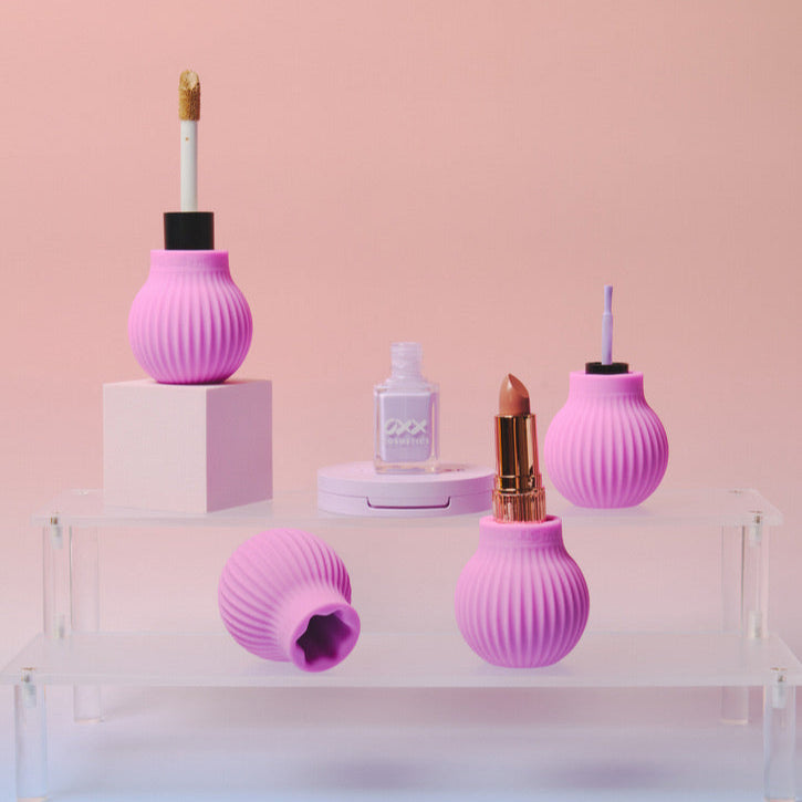 Four pink, ribbed silicone Betty makeup holders by ByStorm, including an ergonomic grip, display lip gloss, nail polish, lipstick, and an empty holder on clear and pink stands against a soft pink background.