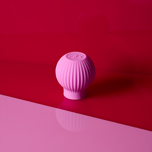 The ByStorm Betty is a pink, round, ribbed ergonomic silicone makeup grip with a textured pattern and raised buttons on top, displayed on a glossy pink and red surface against a red background.