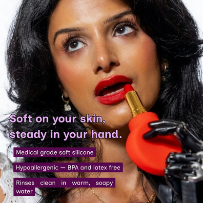 A close-up of a woman applying lipstick with a red paddle-shaped grip. The text reads,  Soft on your skin, steady in your hand, medical-grade soft silicone, hypoallergenic, BPA and latex-free. Rinse clean in warm soapy water. 