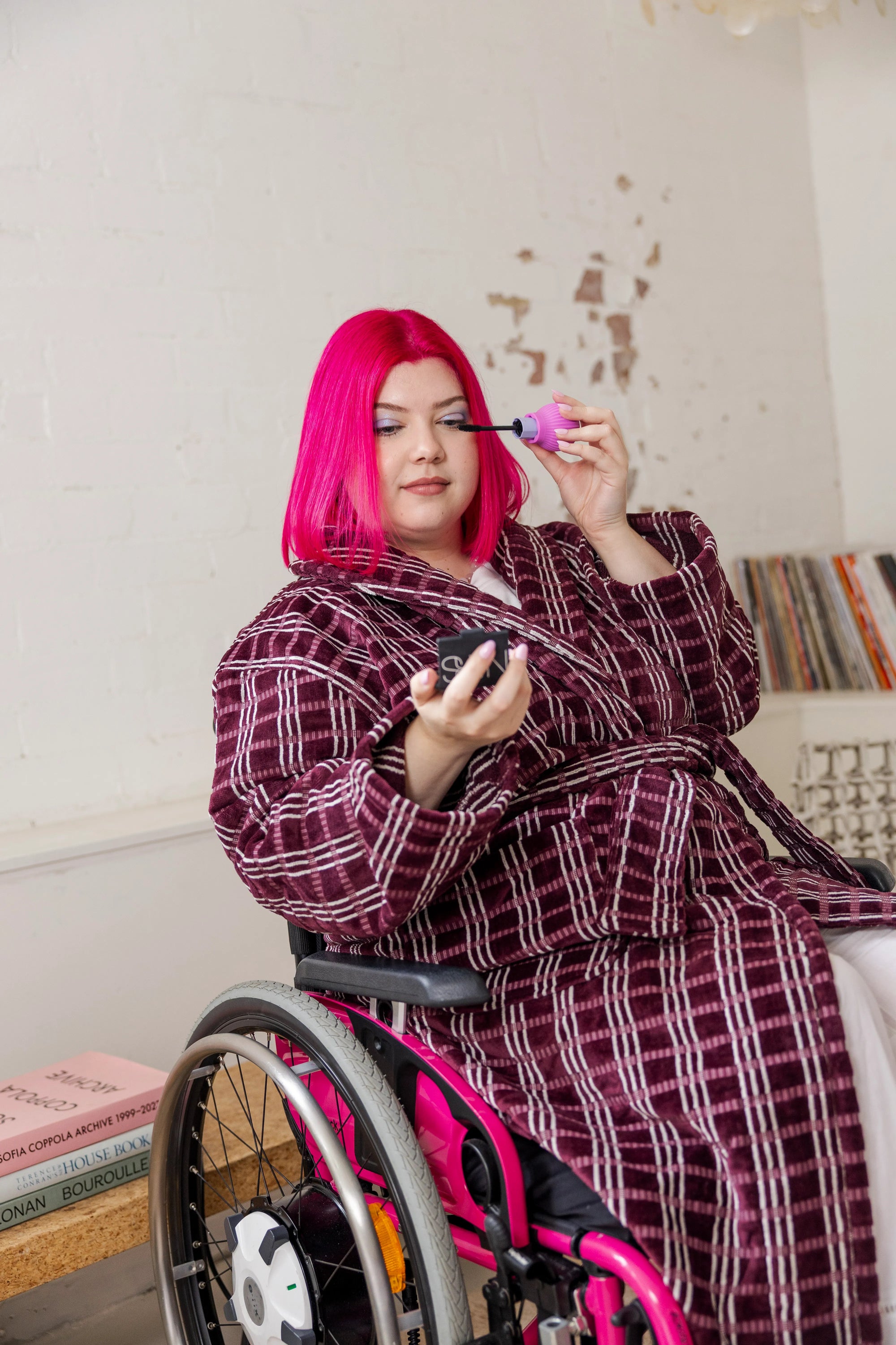 A woman with vibrant pink hair, in a patterned robe, applies makeup in a wheelchair using ByStorm grip tool attached in a mascara. The setting is cozy and artistic, evoking creativity.