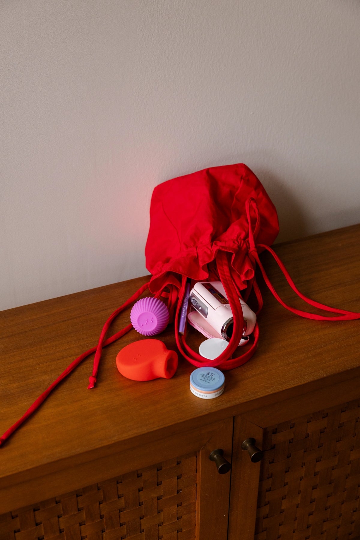 A red drawstring bag is open on a wooden surface, revealing scattered makeup, comb and colorful ByStorm grip tool. The scene is neat, conveying a casual and organized feel.