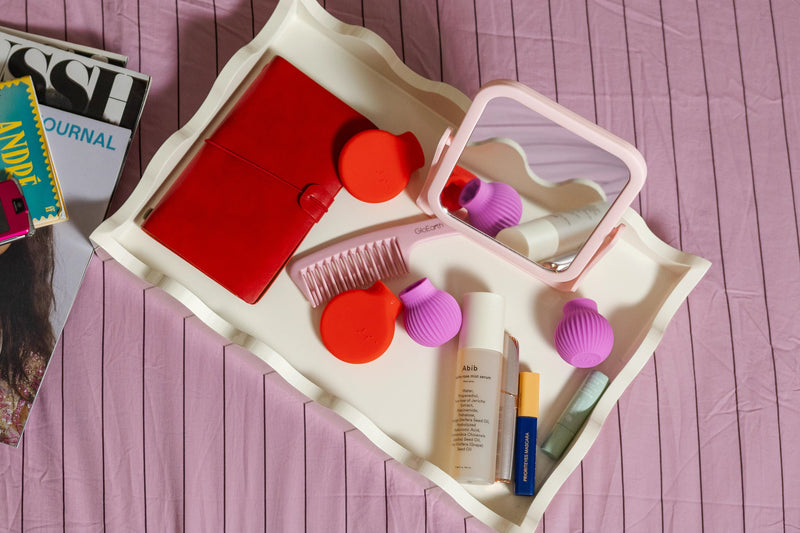 A tray on a pink bedspread holds a red notebook, colorful cosmetics, a mirror, a comb, and ByStorm grip tool in pink and red color. Nearby, fashion magazines and a pink flip phone.