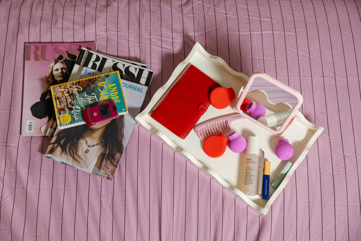 A tray on a pink bedspread holds a red notebook, colorful cosmetics, a mirror, a comb, and ByStorm grip tool in pink and red color. Nearby, fashion magazines and a pink flip phone.
