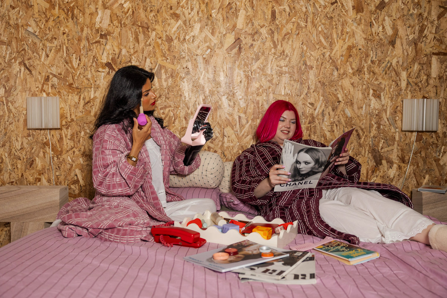 Two people sit on a bed with a pink cover, against a wooden wall. One holds a mirror and ByStorm grip tool applying blush on her cheek, the other reads a fashion magazine. The scene is relaxed.
