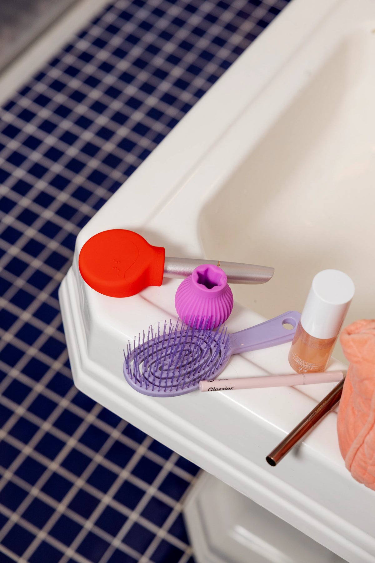 A bathroom scene with a purple hairbrush, red and pink ByStorm grip tool, and a bottle of nail polish on a white bathtub edge. Blue tiled floor in the background.