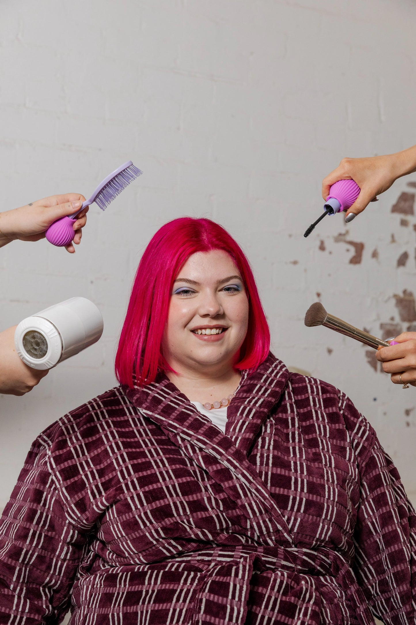 A person with vibrant pink hair smiles while seated wearing a plaid robe. Surrounding hands hold a comb and mascara attached to ByStorm grip tool, hairdryer, and a brush, suggesting a hair makeover.