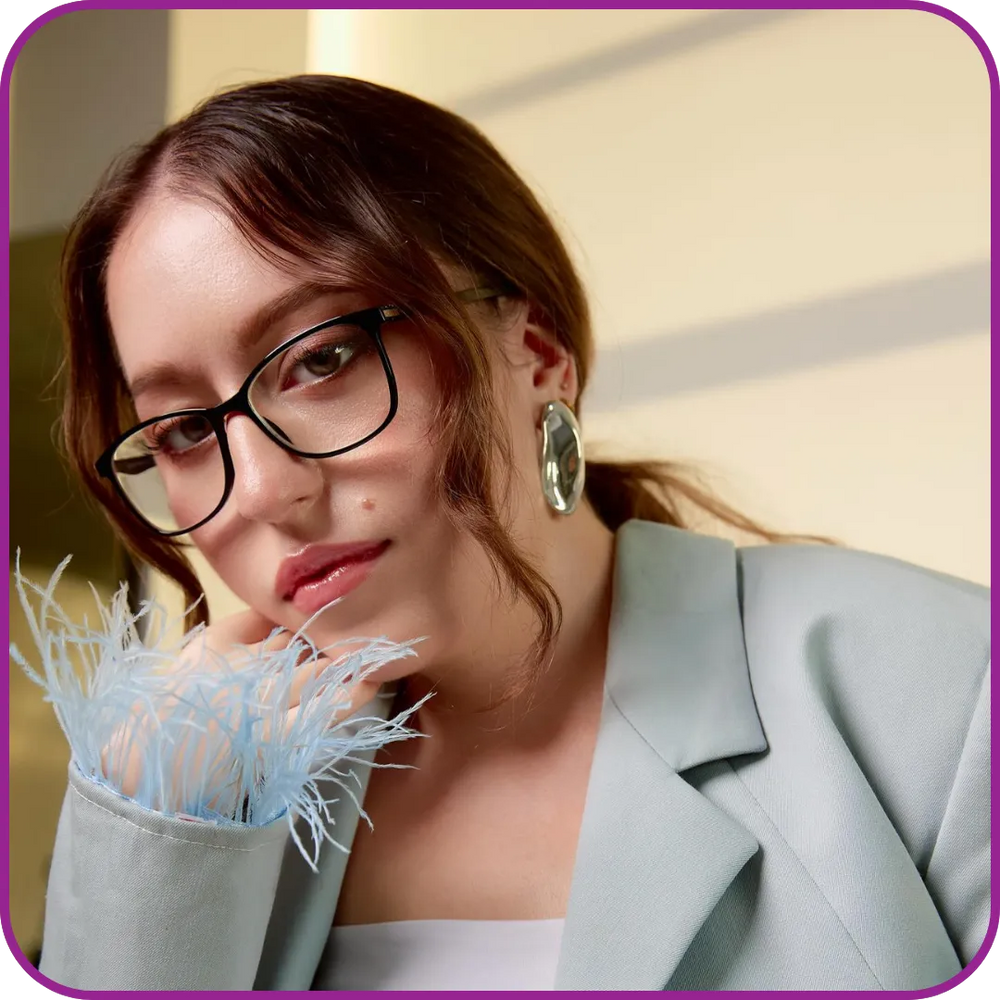 Woman wearing glasses and a light gray blazer with a decorative item in her hand, against a neutral background.