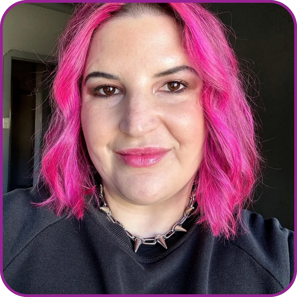 Person with pink hair wearing a black sweater and spiked necklace indoors.