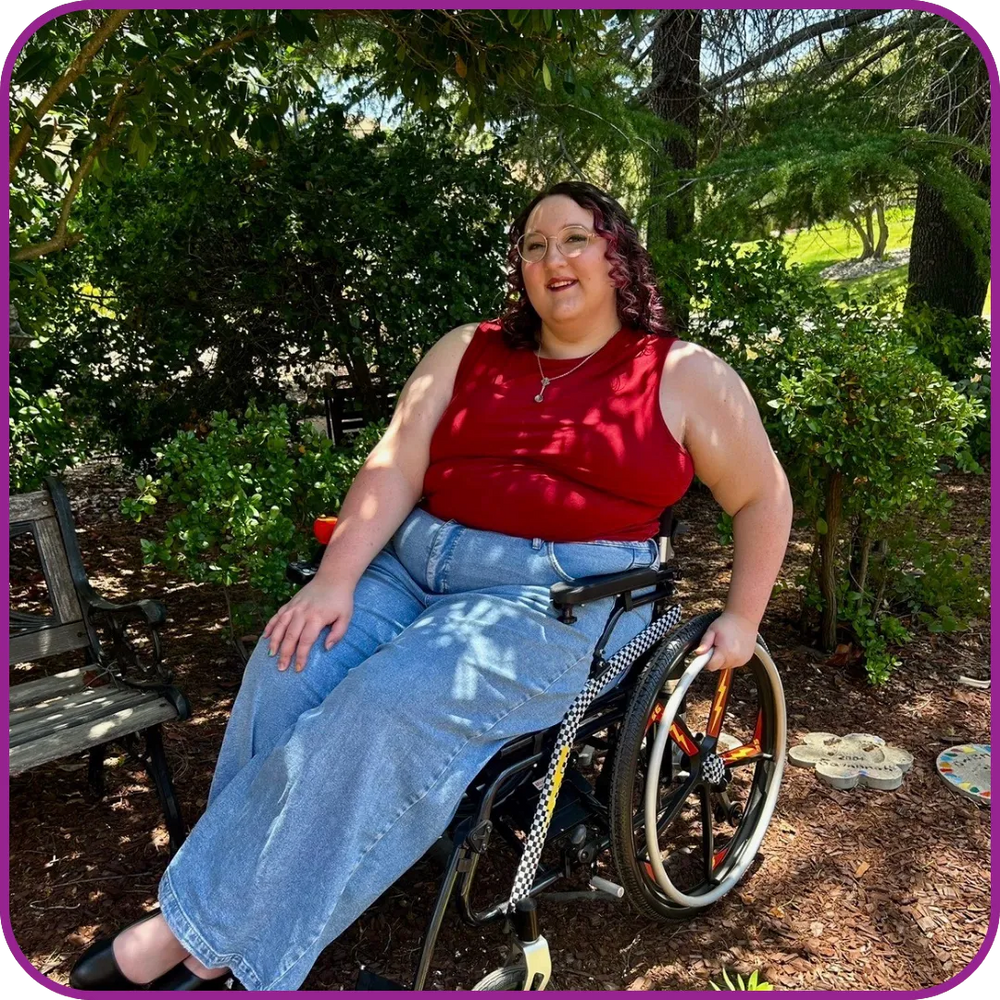 Person in a wheelchair wearing a red top and blue jeans sitting outdoors with greenery around.