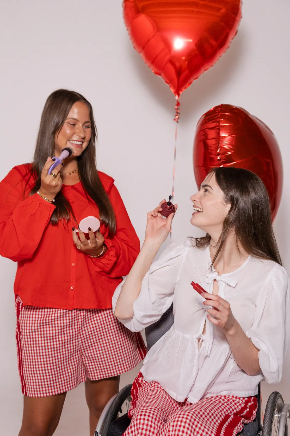 Two disabled women smiling while applying makeup together, holding accessible makeup tools tools, with red heart balloons behind them. 