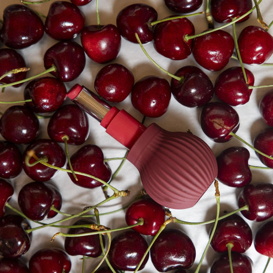 Betty a mauve ball-shaped accessible makeup tool in a lipstick surrounded by cherries on a white background