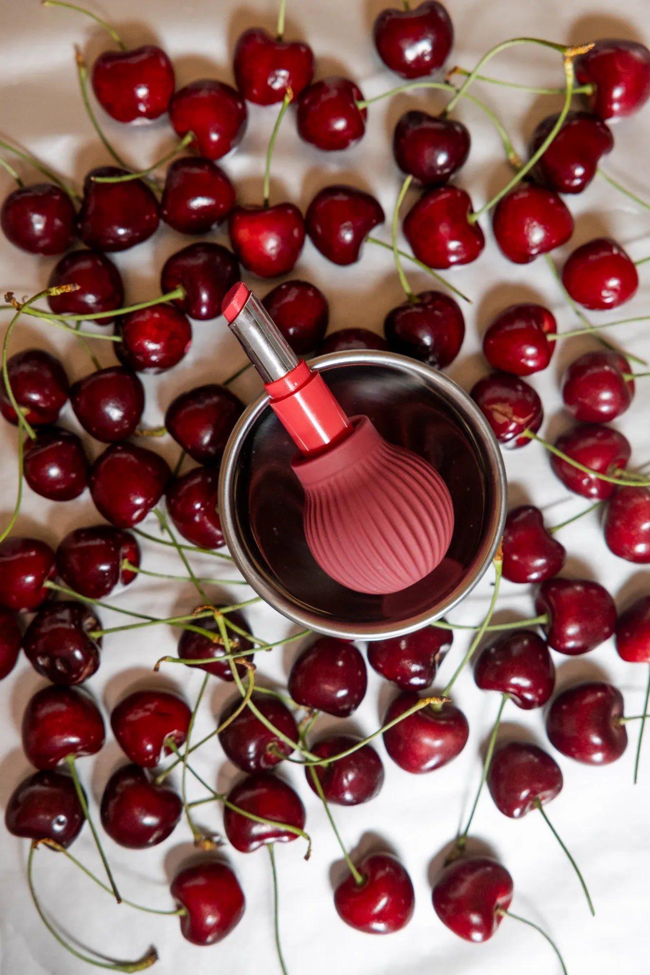 Betty is a mauve ball-shaped accessible makeup tool sits in a bowl surrounded by cherries on a white background