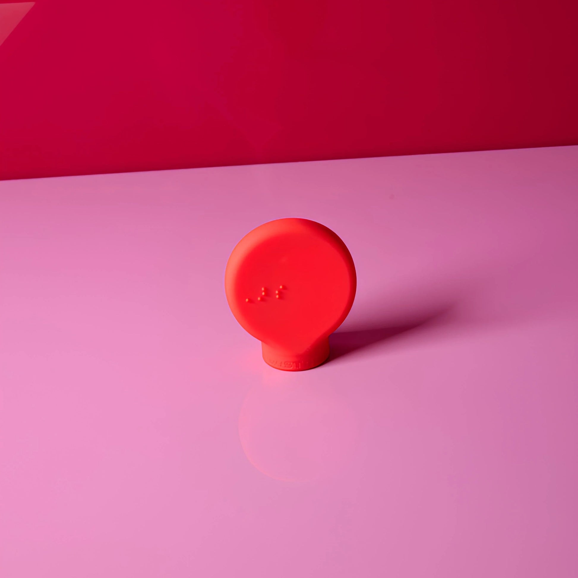 The ByStorm Margie is a small, round, bright red ergonomic beauty tool with a flat base that stands on a smooth, light pink surface against a darker pink background. Subtle raised dots on its front ensure an improved grip.