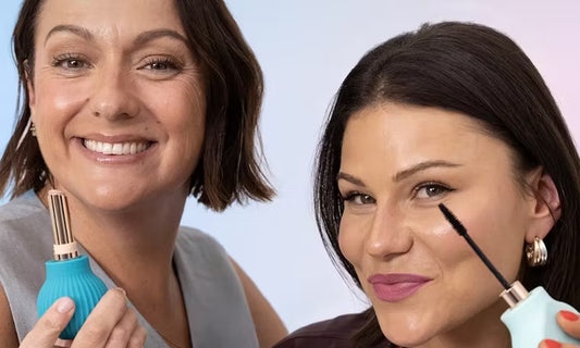 Two women smile at the camera, applying mascara with BOOIE x ByStorm accessible beauty tools by ByStorm. A soft blue and pink gradient background highlights the celebration of inclusive beauty for all.