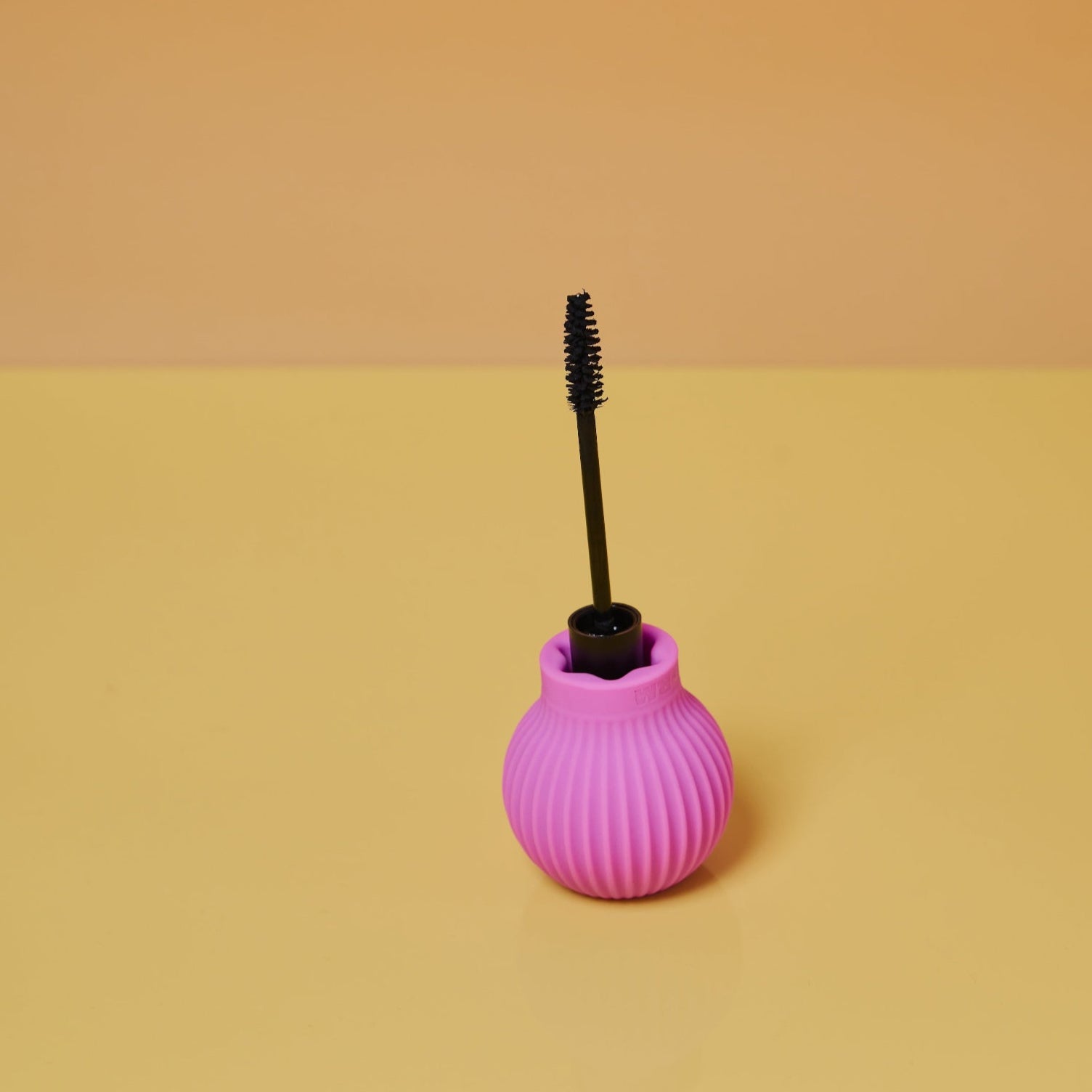 Betty by ByStorm is a pink, ribbed mascara tube with an ergonomic silicone grip, standing upright on a yellow surface with its black wand brush exposed against a pale orange background.