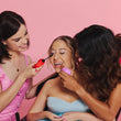 Three women, one in a blue top, smile as they use ByStorm’s Better Together Pack adaptive beauty tools. Against a vibrant pink background, they enjoy accessible makeup and empowering moments together.