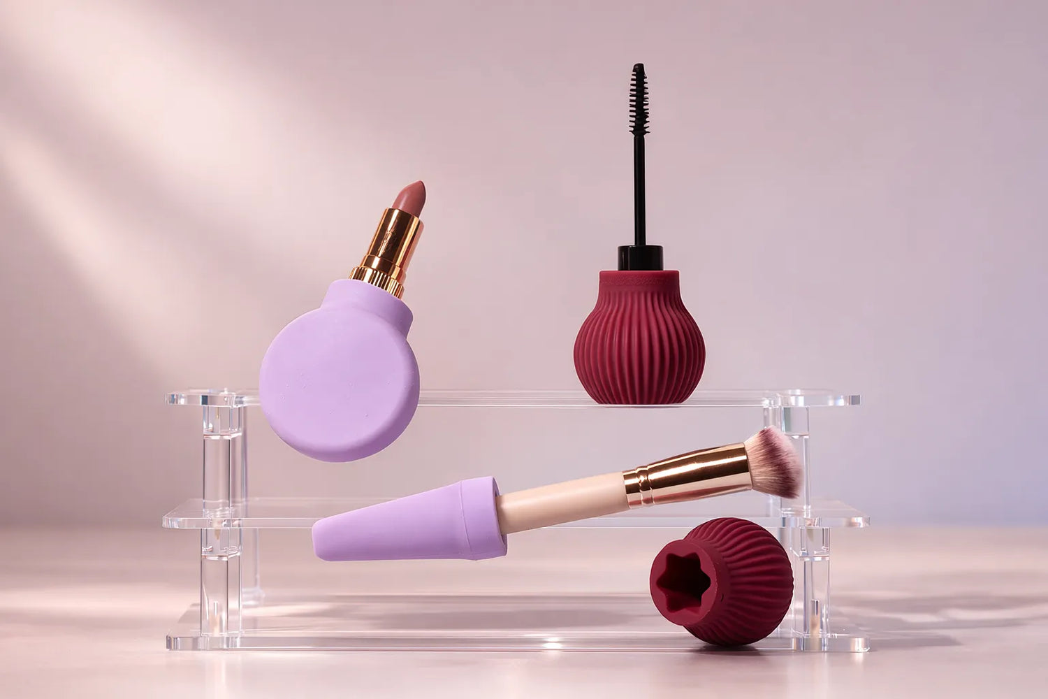 Makeup products including a lip balm, mascara, and brush attached to ByStorm accessible grip tool on a clear stand with a gradient purple background.