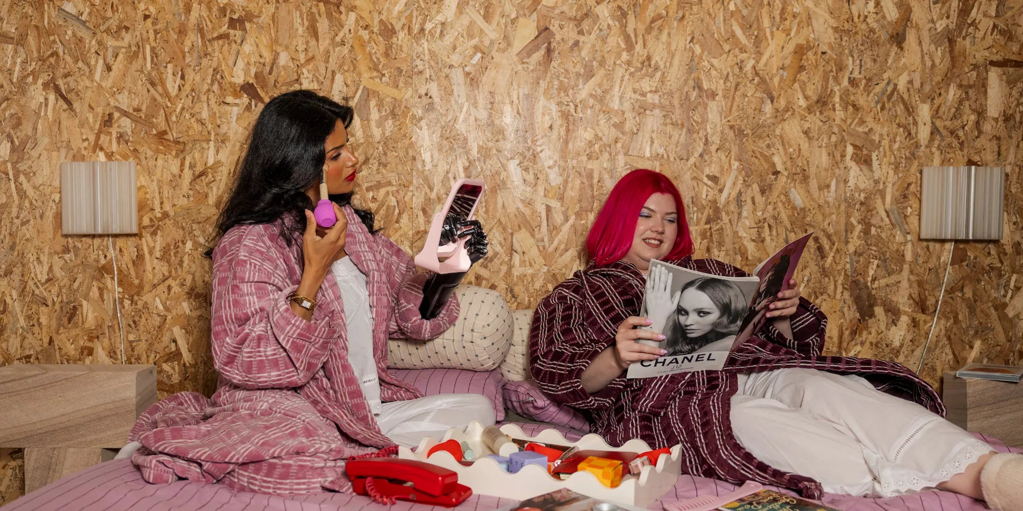 Two models in a cozy bedroom setting, one with black hair holding the ByStorm Adaptive Makeup Grip tool attached to a brush for easier makeup application