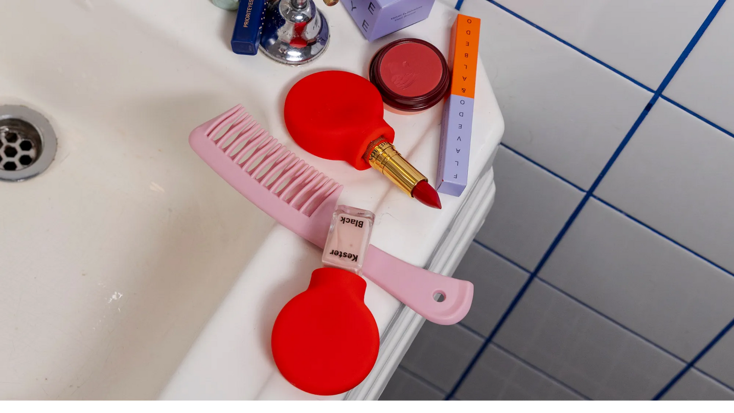 ByStorm Margie red adaptive makeup grip tool styled with a lipstick on a bathroom sink ledge alongside a pink comb and beauty products