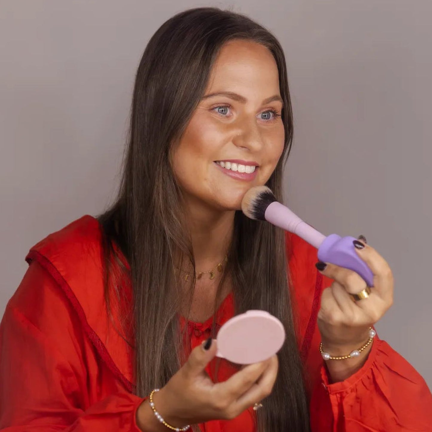 Woman applying blush using Margie, a lilac, flat, paddle-shaped accessible makeup tool of ByStorm for easier application.