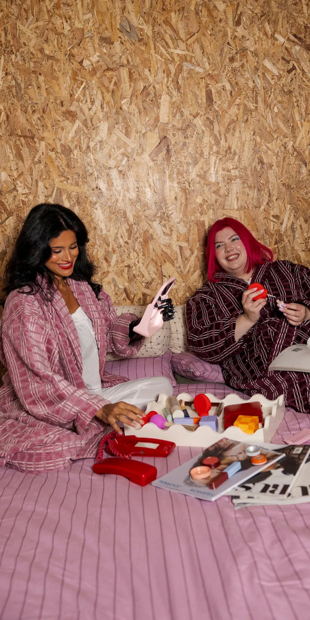 Two models in a cozy bedroom setting, one with pink hair holding the ByStorm Adaptive Makeup Grip tool surrounded by makeup products