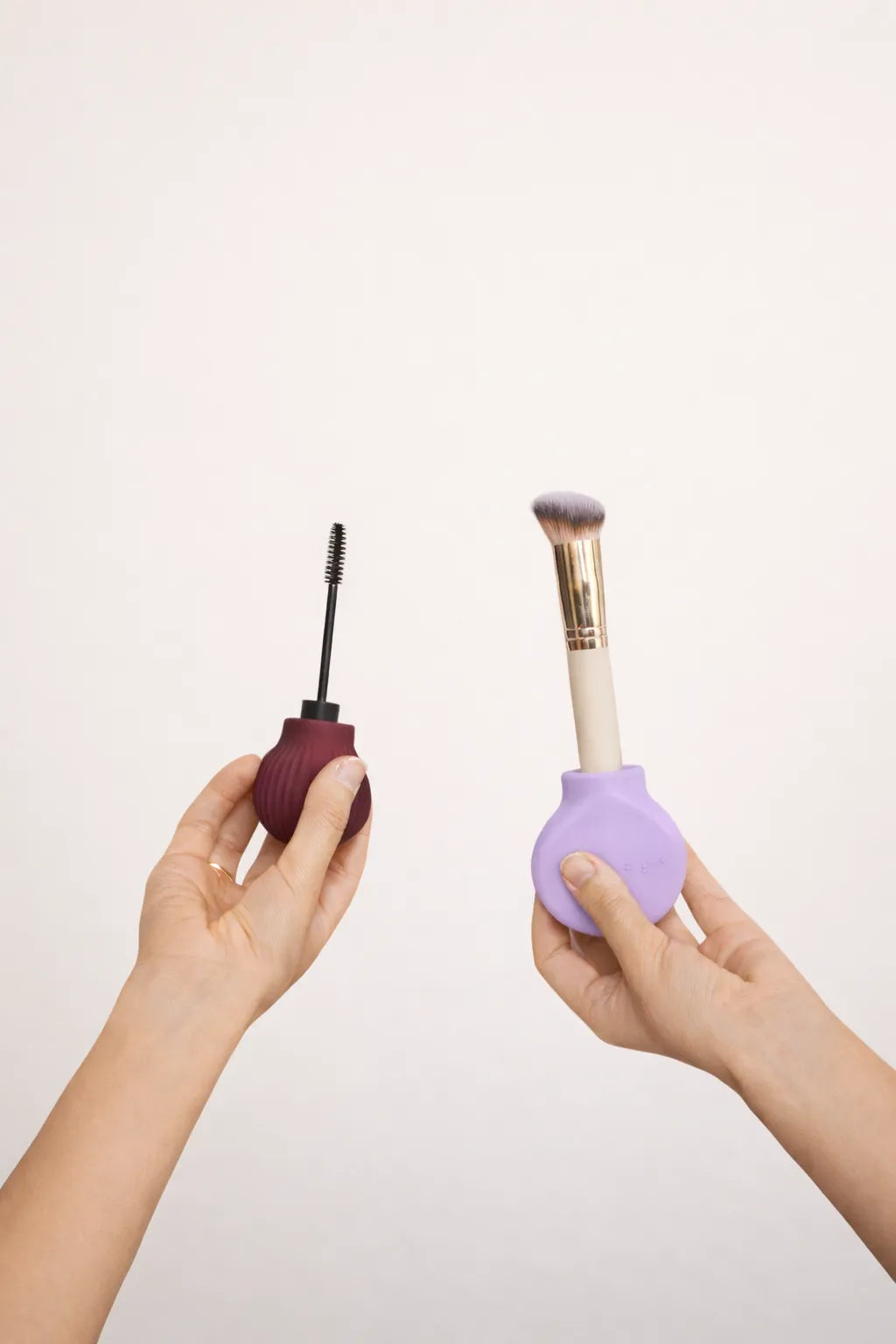 Two hands holding makeup brushes with ByStorm accessible grip tool against a light pink background