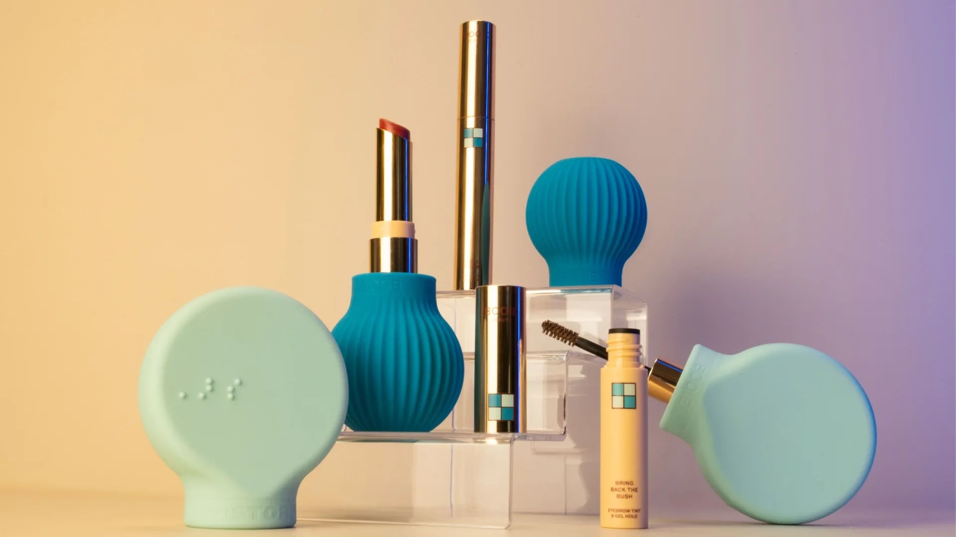 ByStorm’s All The Beauty Items That Landed On Our Desk This Week features teal and gold makeup tubes, blue textured silicone tools, and a yellow mascara tube, all displayed on clear stands against a beige gradient backdrop.