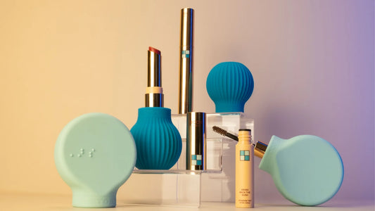 ByStorm’s All The Beauty Items That Landed On Our Desk This Week features teal and gold makeup tubes, blue textured silicone tools, and a yellow mascara tube, all displayed on clear stands against a beige gradient backdrop.