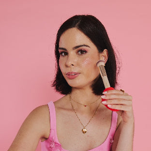 A woman with short dark hair and a pink top applies blush to her cheek using the Margie arthritis-friendly makeup brush by ByStorm, creating a natural look with pink eyeshadow and lipstick against a solid pink background.