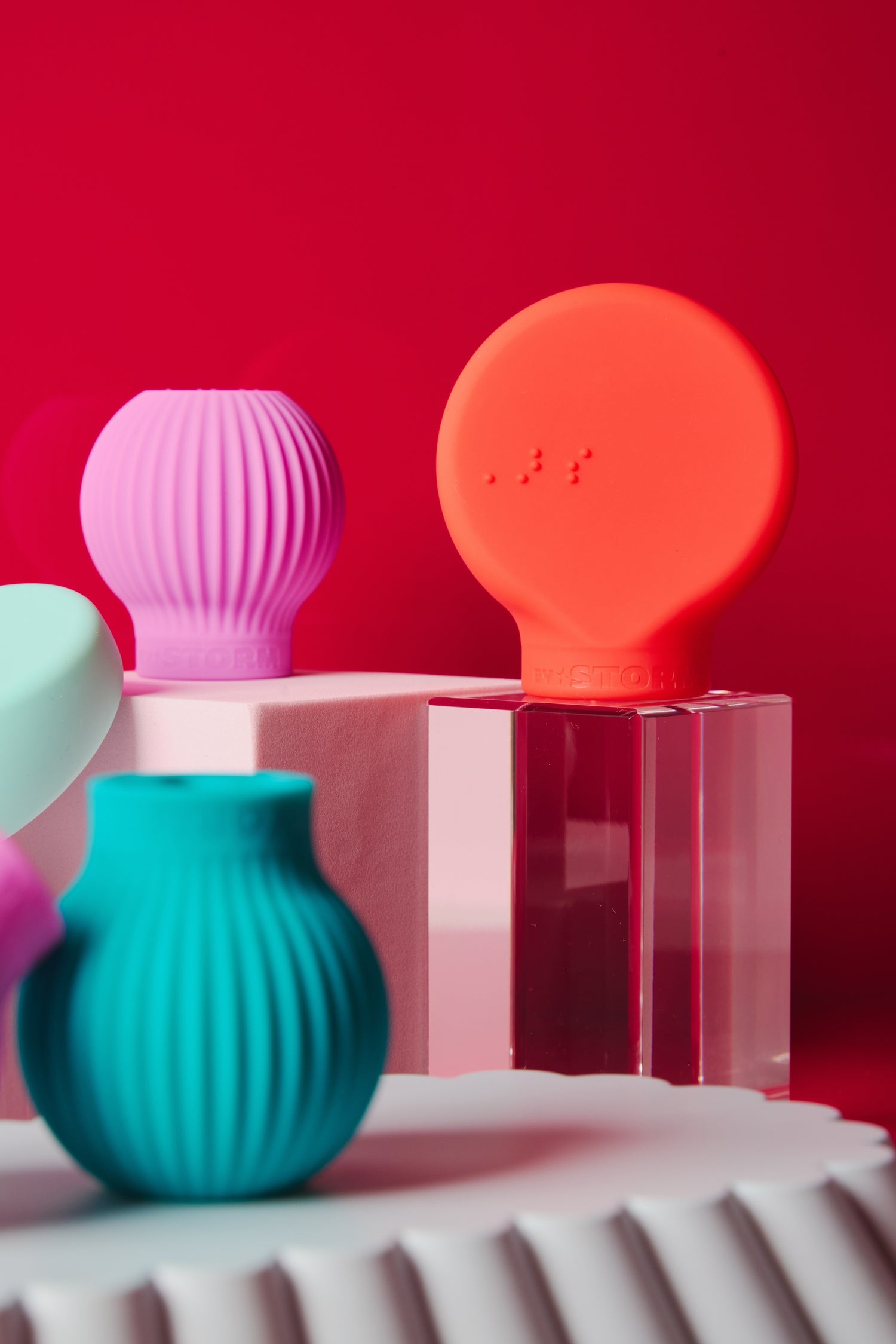 Colorful silicone objects, including round and ribbed shapes in teal, pink, and coral, are displayed on clear and white platforms against a vibrant red background.
