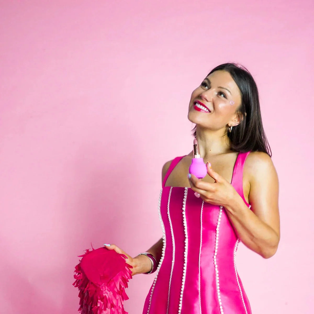 Storm, a women with dark brown hair wearing a hot pink dress on a light pink background holds Betty, a pink ball-shaped adaptive beauty tool holding a lipstick. 